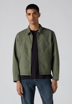 Ung mand iført en grøn lynlåsjakke over en sort trøje og blå jeans, står foran en ensfarvet grå baggrund.