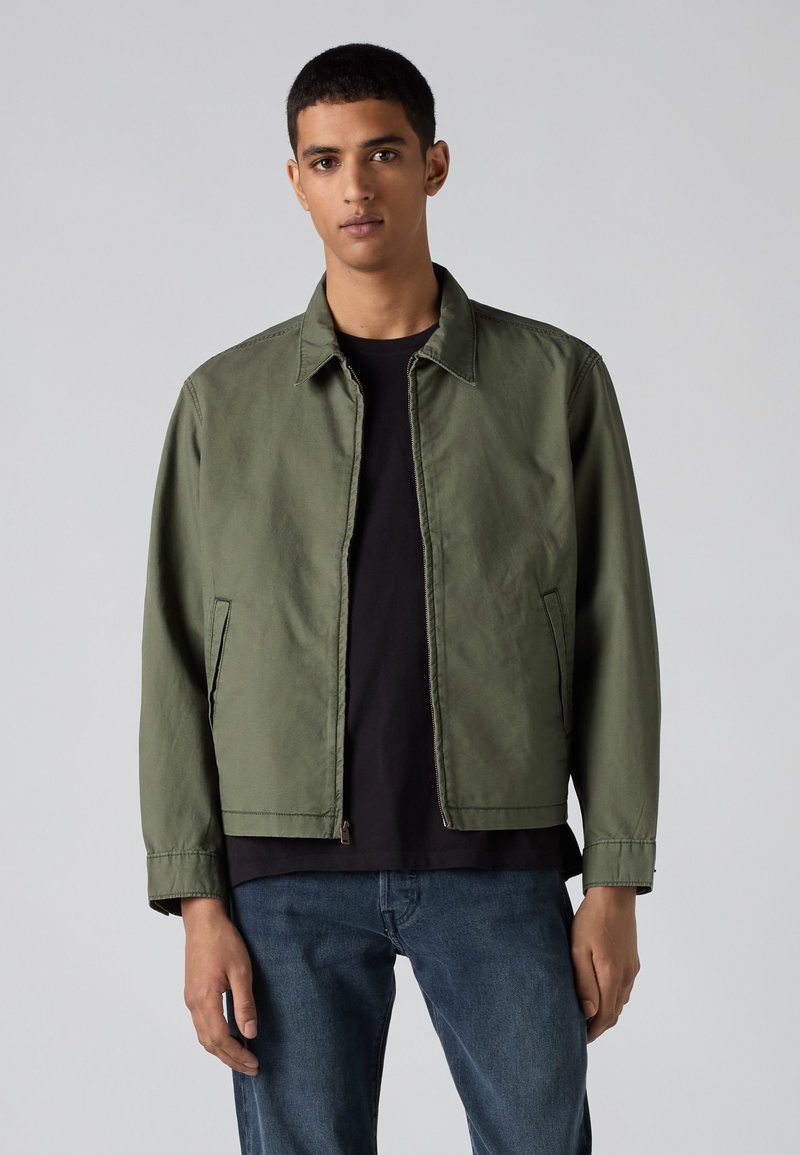 Joven que lleva una chaqueta verde con cremallera sobre una camiseta negra y vaqueros azules, de pie frente a un fondo gris liso.
