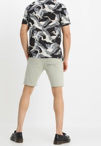 Shorts gris clair avec une texture lisse et une coupe ajustée, associés à une chemise noire à motifs avec des oiseaux blancs. Des chaussures noires complètent le look.