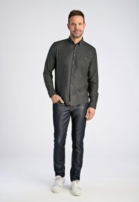 Camisa abotonada de color gris oscuro con un bolsillo, combinada con vaqueros oscuros y zapatillas blancas, sobre un fondo claro. Ajuste casual y textura visible.
