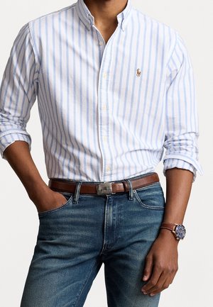 Homme portant une chemise à rayures blanches et bleu clair à manches retroussées, un jean bleu, une ceinture marron et une montre à bracelet en cuir marron.