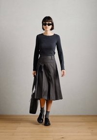 Šedý dlhý rukáv a tmavošedá plisovaná sukňa s vysokým pásom. Čierne slnečné okuliare, tmavé topánky a šedé ponožky. Minimalistický dizajn s čistými líniami.