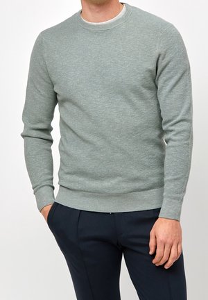Homme portant un pull en maille texturée gris clair à manches longues et un pantalon bleu marine foncé, les mains détendues le long du corps, sur un fond uni.