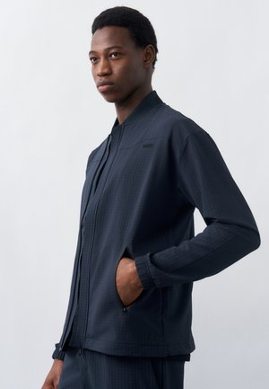 Άνδρας με το χέρι στην τσέπη, φοράει σκούρο μπουφάν με φερμουάρ και ταιριαστό παντελόνι, στρέφεται προς τα αριστερά μπροστά σε απλό φωτεινό φόντο.