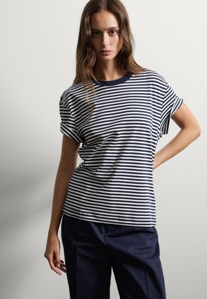 T-shirt a righe blu navy e bianche con maniche corte e colletto rotondo; tessuto morbido; vestibilità rilassata; mostrato con pantaloni scuri; modello in posa con una mano sui fianchi.