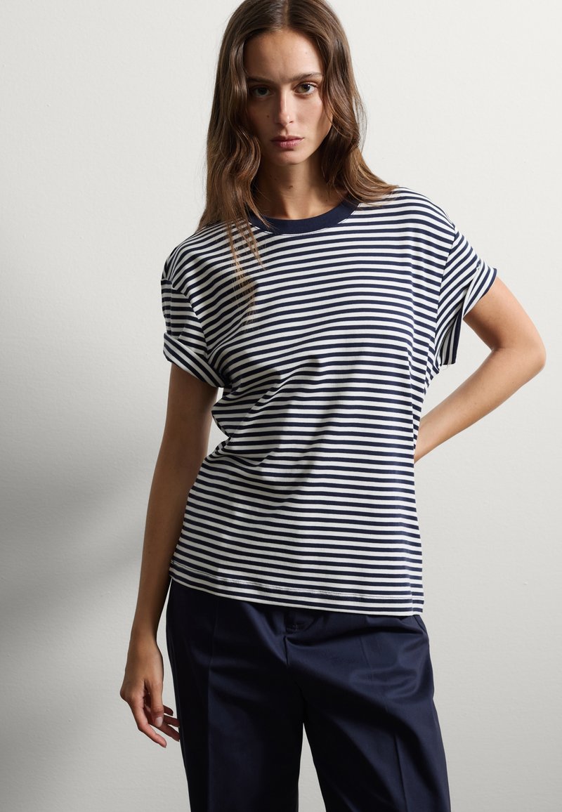 Camiseta de manga corta a rayas en azul marino y blanco con cuello redondo; tejido suave; ajuste relajado; presentada con pantalones oscuros; modelo posando con la mano en la cadera.