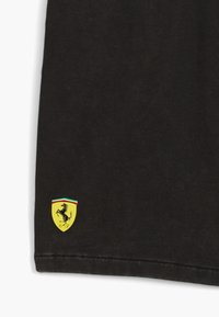 Sorte bomulds T-shirt med et lille gult Ferrari-logo med en springende hest, omkranset af rødt og grønt i nederste hjørne.