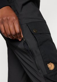 Black cargo pants with a zip pocket, featuring a leather logo patch. Material appears durable with a smooth texture.