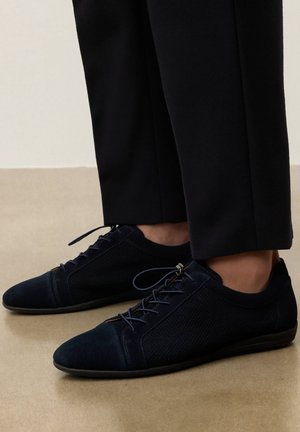 Melni zemo profilu mežģīņu apavi ar acs sāniem, valkāti kopā ar melnām biksēm uz gaiša grīdas seguma.