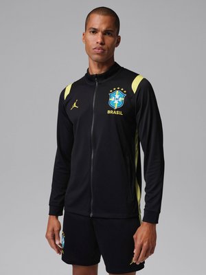 Άνδρας μοντέλο που φοράει μαύρο μπουφάν και σορτς ποδοσφαίρου της Βραζιλίας με κίτρινες λεπτομέρειες, στέκεται μπροστά σε μια απλή γκρι φόντο.