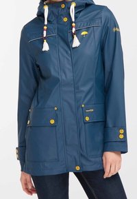 Laivastonsininen vedenpitävä takki hupulla, valkoisilla nyöreillä, joissa on keltaisia ja punaisia helmiä, keltaisia nappia, etutaskuja ja sateenvarjo-logo rintataskussa.