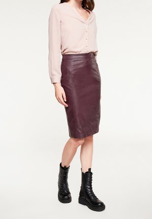 Femme portant un chemisier rose pâle boutonné, rentré dans une jupe en cuir bordeaux longueur genou, avec des bottes de combat noires à lacets.