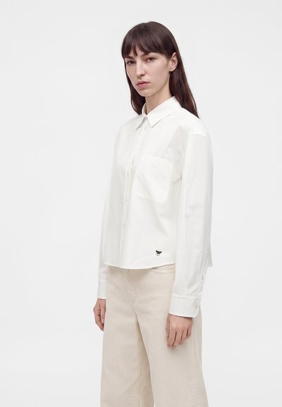Femme aux cheveux bruns et raides portant une chemise blanche à boutons avec une poche avant et un pantalon large beige clair, debout devant un fond uni.