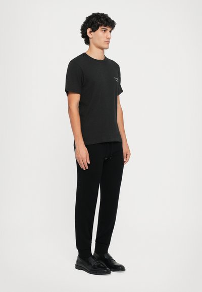 Camiseta negra de manga corta con cuello redondo, combinada con pantalones negros ajustados y zapatos negros sin cordones. Diseño minimalista y textura suave.