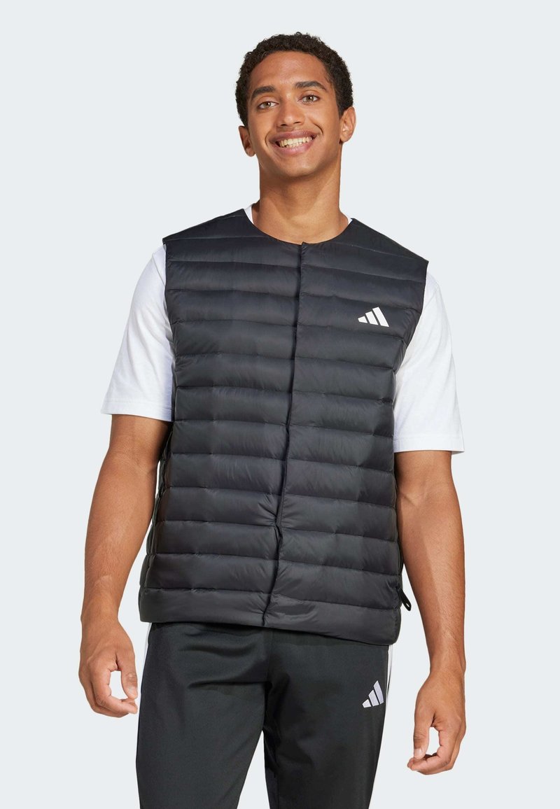 Veste noire matelassée avec un design en surpiqûres, fermeture à boutons-pression sur le devant et logo Adidas blanc sur la poitrine gauche. Portée par-dessus une chemise blanche.