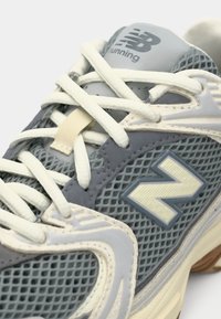 Gros plan d'une chaussure de course New Balance gris et crème avec des panneaux en mesh et des lacets blancs passés dans des œillets.