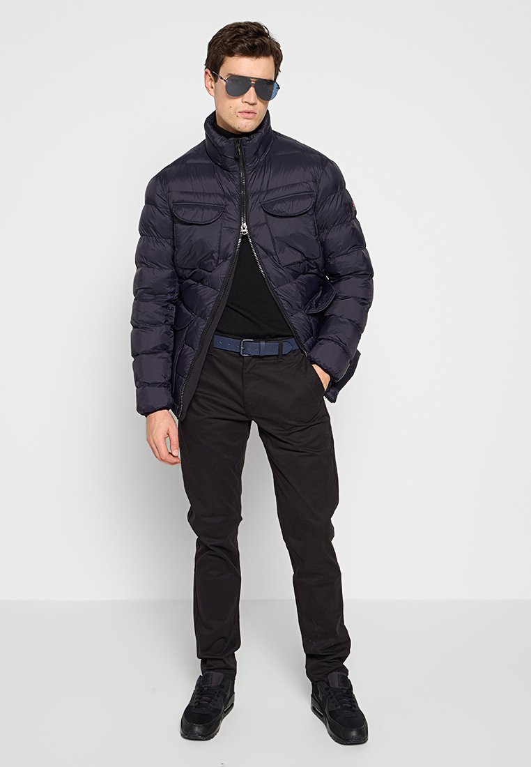 Marineblauwe pufferjack met een rits aan de voorkant, twee borstzakken en een hoge kraag. Draag het over een zwart t-shirt, gecombineerd met zwarte broeken en schoenen.