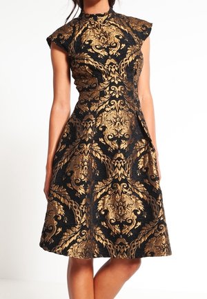 Femme portant une robe noire avec un motif baroque doré orné, col haut, manches courtes et une jupe évasée jusqu'aux genoux.