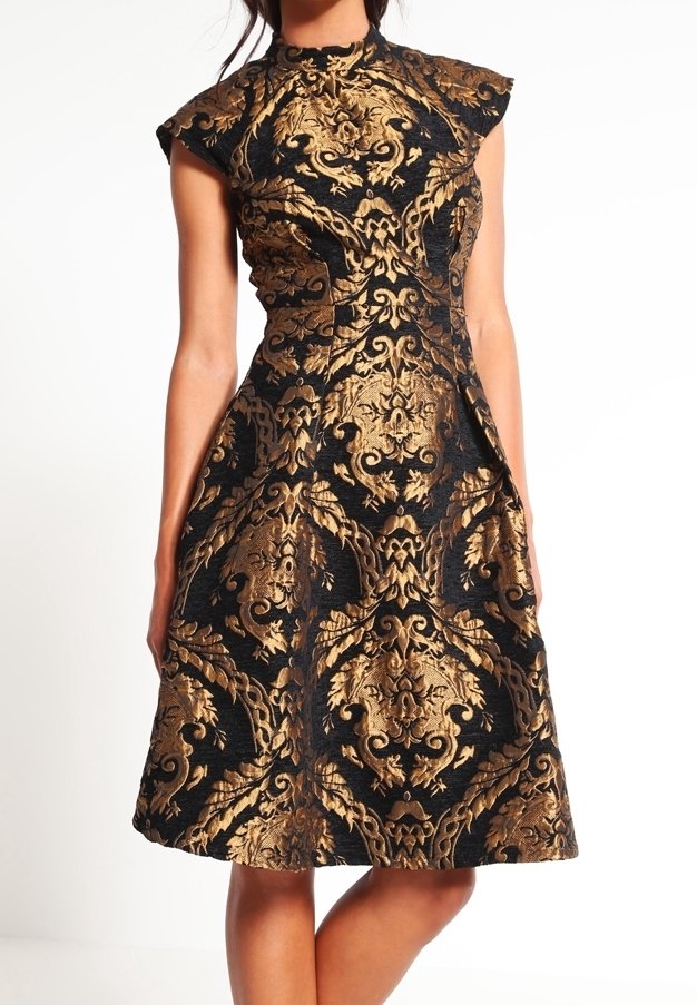 Femme portant une robe noire avec un motif baroque doré orné, col haut, manches courtes et une jupe évasée jusqu'aux genoux.