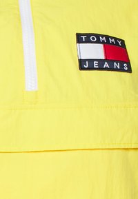 Pullover giallo con zip frontale e un'etichetta nera con la scritta "TOMMY JEANS" e un design rosso, bianco e blu. Tessuto testurizzato.