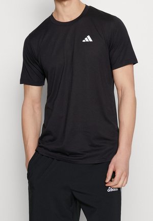 Mann trägt ein schwarzes Adidas T-Shirt und schwarze Adidas-Hose mit weißen Logos, steht mit einer Hand in der Tasche vor einfarbigem Hintergrund.