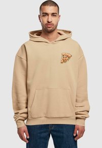 Beige kapucnis pulóver elülső zsebbel, bő szabásban, húzózsinórral ellátott kapucnival, és egy pizza szelet hímzéssel a bal mellen. Az anyag lágy érzetű.