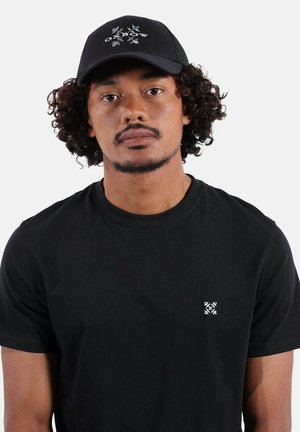 Homme aux cheveux bouclés portant une casquette noire ornée de feuilles bleues et un t-shirt noir avec un petit logo géométrique blanc sur la poitrine.