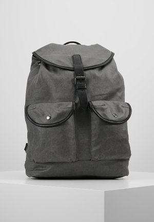 Dunkelgrauer Canvas-Rucksack mit zwei vorderen Klappentaschen und einem oberen Klappdeckel, der durch einen schwarzen Schnallenriemen gesichert ist, steht auf einer weißen Oberfläche.