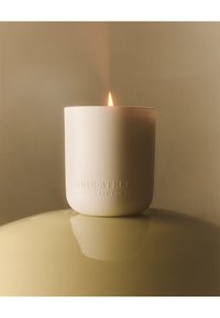 Candelina in ceramica bianca con finitura liscia, caratterizzata da una piccola fiamma. Testo impresso su un lato. Posizionata su uno sfondo lucido e chiaro.