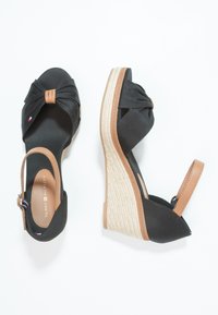 Paire de sandales compensées noires avec brides de cheville couleur camel, devant en tissu noué et texture tissée sur les talons compensés, sur fond blanc.