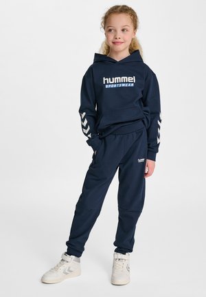 Navyblauw sportkledingset met een sweatshirt met capuchon, voorzien van een logo aan de voorkant en chevronaccenten op de mouwen, gecombineerd met bijpassende joggers en witte sneakers.