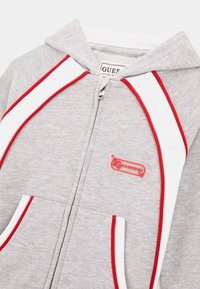 Grauer Reißverschluss-Hoodie mit Kapuze, ausgestattet mit roten und weißen Akzentstreifen, einem auffälligen Logo-Patch und zwei Fronttaschen. Weiche Stofftextur.