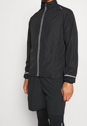 Mann trägt schwarze leichte Jacke mit weißem vertikalem Reißverschluss und passende schwarze Shorts und Leggings vor einfarbigem Hintergrund.