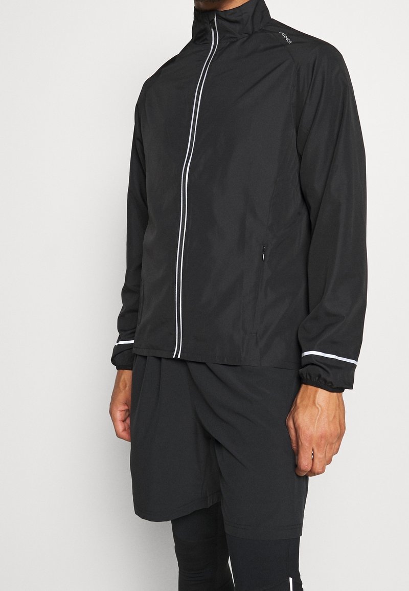 Homme portant une veste légère noire avec une fermeture éclair verticale blanche et un short noir assorti ainsi que des leggings sur un fond uni.