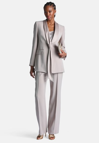 Femme portant un tailleur pantalon gris clair avec un blazer à col châle noué, tenant une pochette beige, et des talons ouverts dorés.