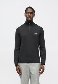 Camisola preta com fecho de correr no pescoço, confeccionada em tecido de malha macio, com detalhes canelados e um pequeno logótipo branco no peito. Usada com calças cinzentas às riscas.