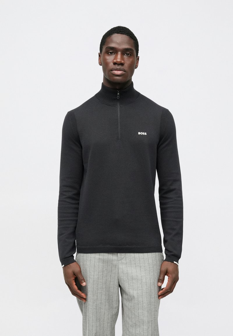 Camisola preta com fecho de correr no pescoço, confeccionada em tecido de malha macio, com detalhes canelados e um pequeno logótipo branco no peito. Usada com calças cinzentas às riscas.