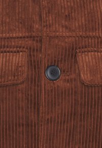 Brun kordflorsliknande tyg med vertikala ränder, med en enda svart knapp och två påsydda fickor fram. Strukturerad yta.