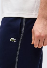 Κοντινή λήψη ενός ατόμου που φοράει μπλε σκούρο παντελόνι Lacoste με λευκές πλαϊνές ρίγες με το λογότυπο και ένα λευκό πουκάμισο, με το χέρι χαλαρό δίπλα στο σώμα.