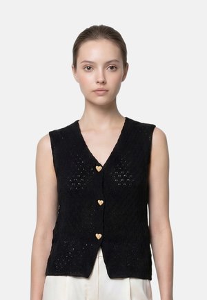 Giovane donna con capelli castani raccolti indossa un cardigan nero senza maniche con tre bottoni a forma di cuore e pantaloni bianchi.