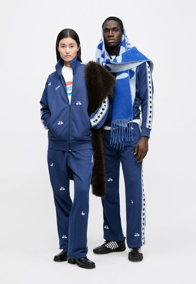 Navy tracksuits with white embroidered bunny logos, featuring stripes and stars on the sides. The male model drapes a blue and white patterned scarf.