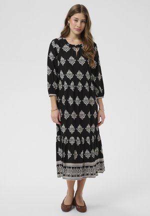 Jeune femme debout portant une robe midi noire à motifs floraux blancs avec des manches trois-quarts, associée à des chaussures plates tressées marron.