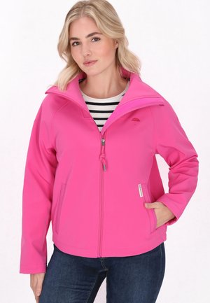 Kvinde med blondt hår iført en knald pink lynlåstrøje over en sort-hvidstribet skjorte og mørke jeans, den ene hånd i jakke lommen.