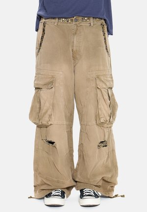 Pantaloni cargo beige a gamba larga con vita borchiata, ginocchia strappate, grandi tasche laterali, abbinati a sneakers nere e bianche.