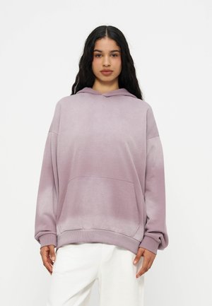 Femme aux longs cheveux bruns portant un sweat à capuche ample violet clair et un pantalon blanc, debout devant un fond blanc uni.