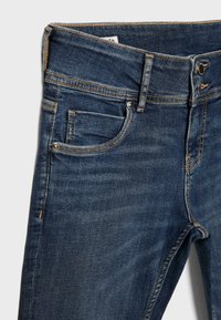 Jean en denim bleu foncé présentant des poches avant, des passants pour ceinture, une fermeture par bouton et des coutures contrastées sur un fond clair.