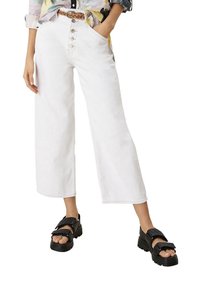 Valkoiset leveälahkeiset housut, joissa on korkea vyötärö ja napillinen edusta, teksturoidusta kankaasta ja suora siluetti. Yhdistetty paksupohjaisiin mustiin sandaaleihin.