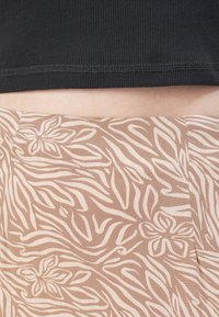 Top negro con canalé y un dobladillo corto sobre una tela marrón clara que presenta un patrón floral y ondulado en tonos crema. Detalle de superficie texturizada visible.