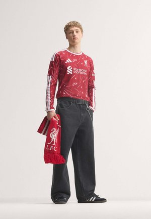 Männlicher Sportler trägt ein rotes Liverpool FC Langarmtrikot und Shorts, steht mit den Händen in der Hüfte und schaut zur Seite.