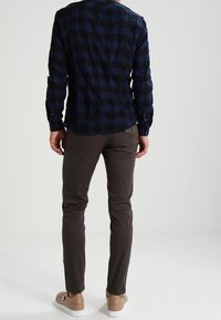 Mies seisoo selin, yllään sinimusta ruudullinen paita, tummanruskeat housut ja beiget tennarit valkoista taustaa vasten.